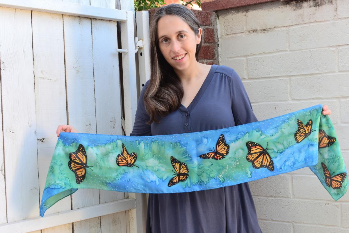 A person with pale skin and long brown hair holds a blue and green scarf decorated with butterflies