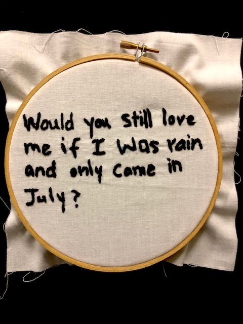 "Would you still love me if I was rain and only came in July?" in black thread on a white canvas background, set in an embroidery hoop.