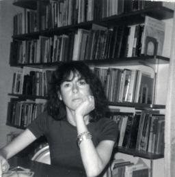 Louise Glück sits at a table with her hand under her chin in front of a bookshelf
