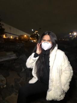 A brown trans woman stands by rocks at night. She is wearing black & a white coat. She is masked and putting her hair behind her ear. In the background are blurry lights from buildings. 