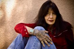 Joy Harjo sits with her knees to her chest and her arm draped over her legs, looking into the camera
