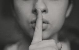 A person holds their pointer finger up to their lips as if to shush someone. Greyscale.