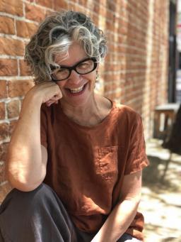 A person with curly gray hair and an ochre tee shirt on sits smiling in front of a brick wall