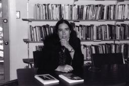 Rebecca Sieferle sits at a table with a hand to her chin in front of bookshelves