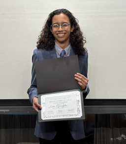 A young person in a suit with long curly hair holds a certificate