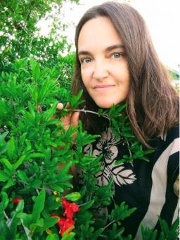 A pale skinned person with long brown hair crouches next to a plant