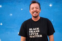 TC Tolbert smiles, wearing a "Black Trans Lives Matter" shirt, standing against a blue wall