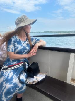 Pale woman with long hair in blue patterned dress and hat looks at the ocean from a boat