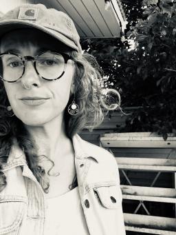 Person with a hat, glasses, and medium length hair stands on a porch