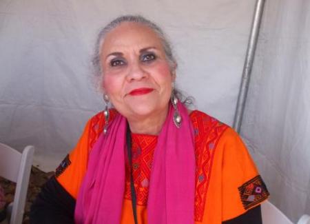 A woman with pulled back gray hair wears a pink and orange outfit in front of a plain background