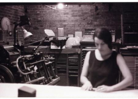A black and white photograph of a woman standing in front of a windmill printing press.