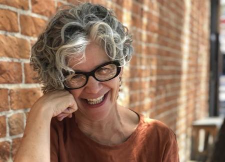 A person with curly gray hair and an ochre tee shirt on sits smiling in front of a brick wall