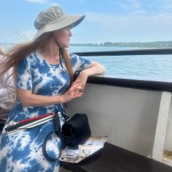 Pale woman with long hair in blue patterned dress and hat looks at the ocean from a boat